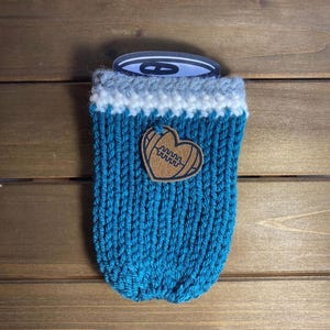 Op de afbeelding: Een turquoise gebreide blikjeshoes met een witte en grijze rand. Een bruin leren hartvormig embleem met een voetbalontwerp is aan de voorkant bevestigd. De hoes is ontworpen om dranken koud te houden.