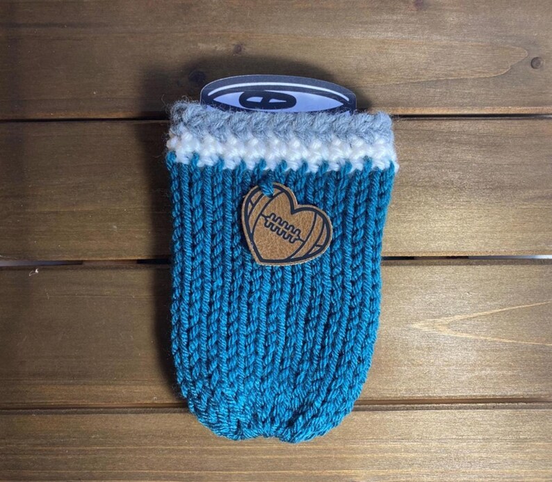 Op de afbeelding: Een turquoise gebreide blikjeshoes met een witte en grijze rand. Een bruin leren hartvormig embleem met een voetbalontwerp is aan de voorkant bevestigd. De hoes is ontworpen om dranken koud te houden.