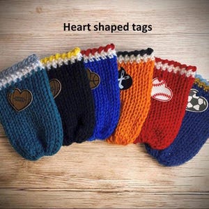 Op de afbeelding: Een verzameling hartvormige labels in verschillende kleuren, waaronder teal, marineblauw, blauw, oranje en rood. Elk label heeft een ander sportthema, zoals een basketbal, honkbal en voetbal. De labels zijn gebreid en hebben een decoratieve rand.