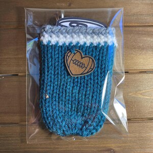 Op de afbeelding: Een tealblauwe gebreide drankhouder met een witte en grijze rand. Een bruine leren hartvormige patch met een voetbalontwerp is aan de voorkant bevestigd. De houder is verpakt in een doorzichtige plastic zak.