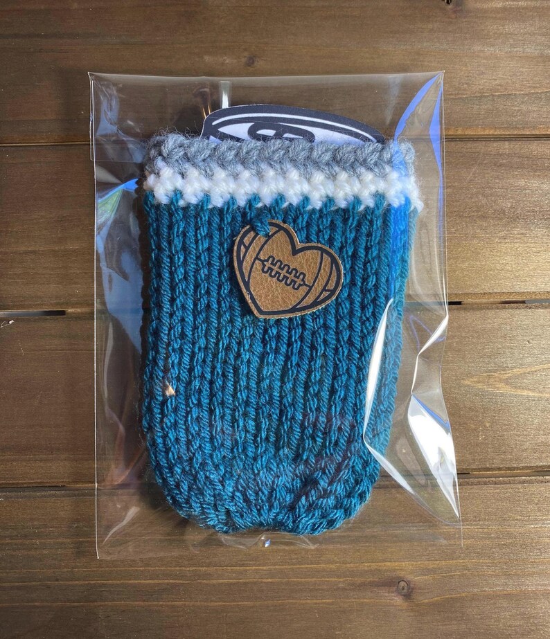 Op de afbeelding: Een tealblauwe gebreide drankhouder met een witte en grijze rand. Een bruine leren hartvormige patch met een voetbalontwerp is aan de voorkant bevestigd. De houder is verpakt in een doorzichtige plastic zak.