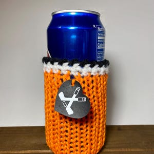 Op de afbeelding: Een oranje gebreide blikjeshouder met een zwarte, witte en oranje rand. Een zwart hartvormig label met gekruiste hockeysticks hangt aan de houder. Een blauw blikje zit erin.