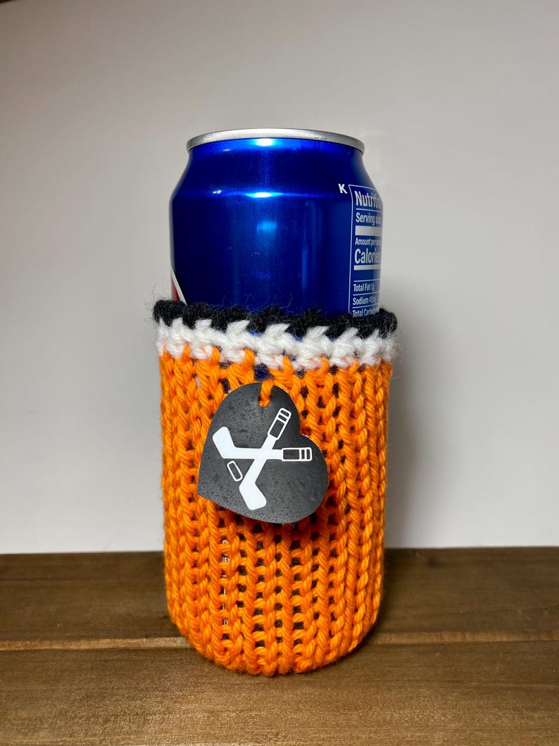 Op de afbeelding: Een oranje gebreide blikjeshouder met een zwarte, witte en oranje rand. Een zwart hartvormig label met gekruiste hockeysticks hangt aan de houder. Een blauw blikje zit erin.