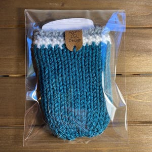 Op de afbeelding: Een turquoise gebreide koffiekopmouw met een witte en grijze rand. De mouw is verpakt in een doorzichtige plastic zak. Een klein bruin label met de tekst "LGL Design" is bevestigd.