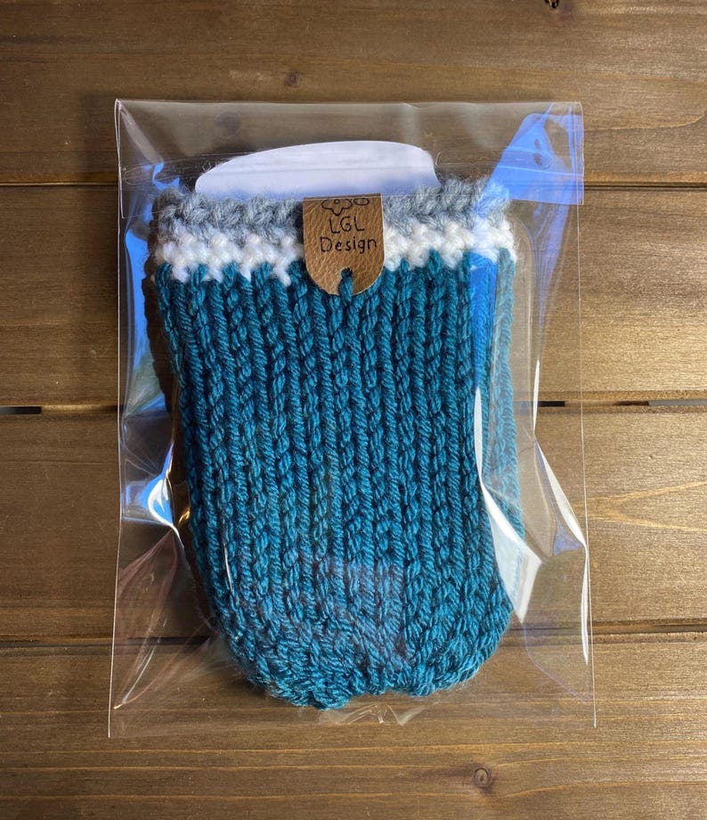 Op de afbeelding: Een turquoise gebreide koffiekopmouw met een witte en grijze rand. De mouw is verpakt in een doorzichtige plastic zak. Een klein bruin label met de tekst "LGL Design" is bevestigd.