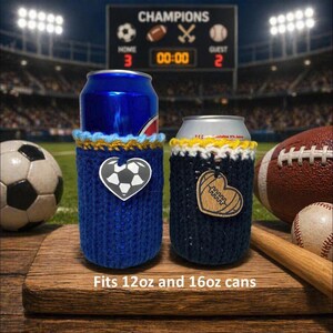 Op de afbeelding: Twee gebreide blikjeskoelers, &eacute;&eacute;n blauw met een voetbalhart, de andere marineblauw met een footballhart. De tekst "Fits 12oz and 16oz cans" staat onderaan. Een voetbal, een rugbybal en een honkbal staan op de achtergrond.