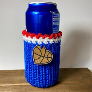 Op de afbeelding: Een blauwe gebreide blikjeshouder met rode en witte rand. Een houten hartvormig label met een basketbalontwerp is bevestigd. Ontworpen om een standaard formaat drankblikje te bevatten.