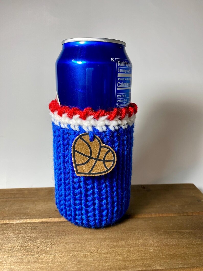 Op de afbeelding: Een blauwe gebreide blikjeshouder met rode en witte rand. Een houten hartvormig label met een basketbalontwerp is bevestigd. Ontworpen om een standaard formaat drankblikje te bevatten.