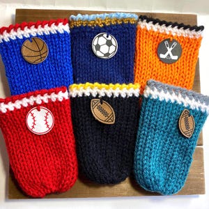 Op de afbeelding: Zes gebreide drankhoezen in verschillende kleuren, elk met een houten embleem met sportthema. De hoezen zijn blauw, rood, marineblauw, oranje en blauwgroen, met witte en gekleurde randen. Emblemen zijn onder meer een basketbal, honkbal, voetbal, hockeysticks en American football.