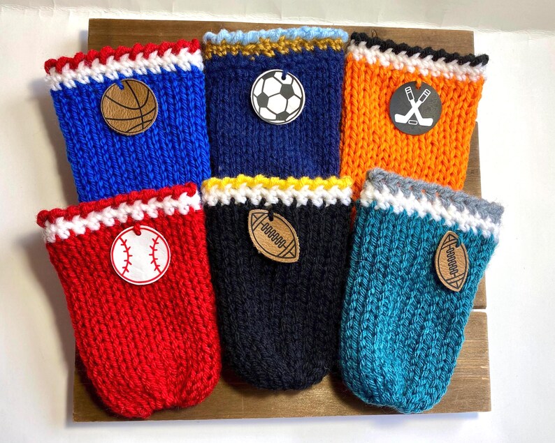 Op de afbeelding: Zes gebreide drankhoezen in verschillende kleuren, elk met een houten embleem met sportthema. De hoezen zijn blauw, rood, marineblauw, oranje en blauwgroen, met witte en gekleurde randen. Emblemen zijn onder meer een basketbal, honkbal, voetbal, hockeysticks en American football.