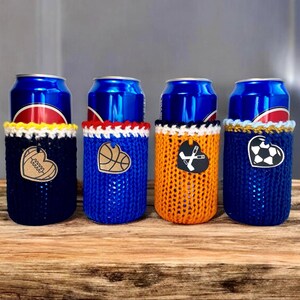 Op de afbeelding: Vier drankblikjeskoelers in marineblauw, blauw, oranje en marineblauw. Elke koeler heeft een ander sportthema hartontwerp: een honkbal, basketbal, hockeysticks en een voetbal. De koelers staan op een houten oppervlak.