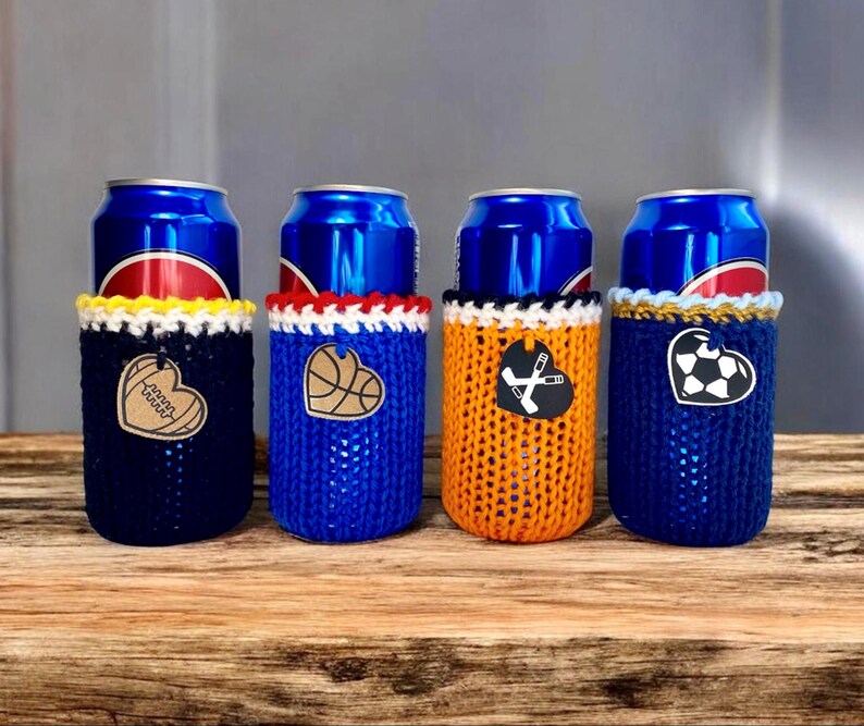 Op de afbeelding: Vier drankblikjeskoelers in marineblauw, blauw, oranje en marineblauw. Elke koeler heeft een ander sportthema hartontwerp: een honkbal, basketbal, hockeysticks en een voetbal. De koelers staan op een houten oppervlak.