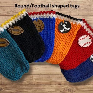 Op de afbeelding: Een verzameling gebreide houders in verschillende kleuren, elk met een sportthema-label. De houders zijn teal, zwart, blauw, oranje, rood en blauw, met labels met een voetbal, basketbal, hockeysticks, honkbal en voetbal.