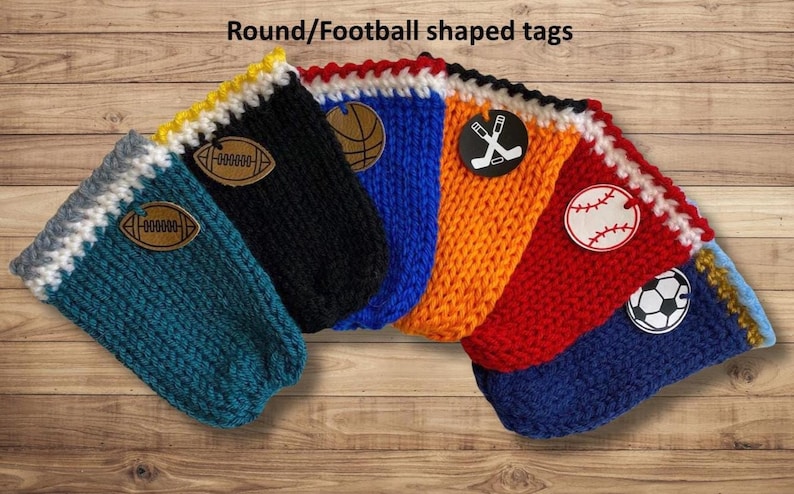 Op de afbeelding: Een verzameling gebreide houders in verschillende kleuren, elk met een sportthema-label. De houders zijn teal, zwart, blauw, oranje, rood en blauw, met labels met een voetbal, basketbal, hockeysticks, honkbal en voetbal.