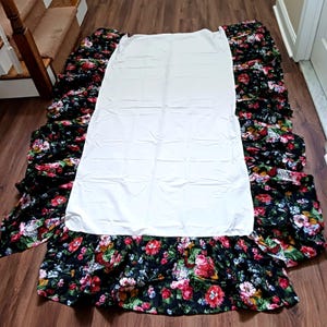 May include: A white bed skirt and matching white sheet are displayed on a wooden floor. The bed skirt has a black floral pattern with red, pink, and yellow flowers. The sheet is plain white.