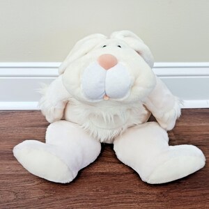 Può includere: Un peluche a forma di coniglio bianco sporco con grandi orecchie, un naso rosa e guance bianche e soffici. Il coniglio è seduto su un pavimento di legno, con le zampe grandi distese. Il giocattolo ha un aspetto morbido e coccoloso.