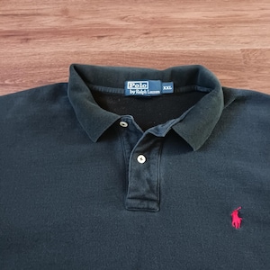 May include: A dark navy blue Polo shirt by Ralph Lauren, size XXL. The shirt features a classic polo collar, a three-button placket, and a small red embroidered polo player logo on the chest. The label reads "Polo by Ralph Lauren".