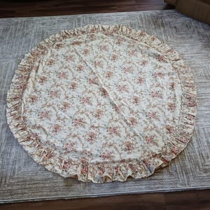 May include: A circular fabric tree skirt featuring a cream-colored base with a repeating floral pattern in shades of pink, red, and green. The edge of the circular fabric is finished with a gathered, ruffled border in the same floral print.