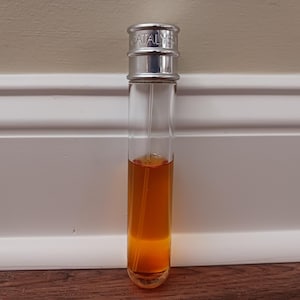May include: A tall, cylindrical glass perfume bottle filled with an amber-coloured liquid. The bottle features a metallic silver cap with the word CATALYST embossed on the top. A thin, clear plastic spray tube is visible inside the glass container.