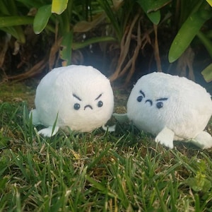 Könnte beinhalten: Zwei weiße Plüschtiere mit wütendem Gesichtsausdruck und kleinen Armen und Beinen. Die Spielzeuge sitzen auf einem Bett aus grünem Gras.