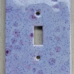 May include: A decorative light switch cover with a glossy, speckled finish. The cover is primarily light purple with darker purple and black speckles and floral-like patterns. It has a rectangular opening for the switch and two holes for mounting.