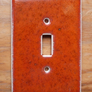 May include: A ceramic light switch cover with a textured orange glaze. The switch cover has a white border and a rectangular cutout for the switch.