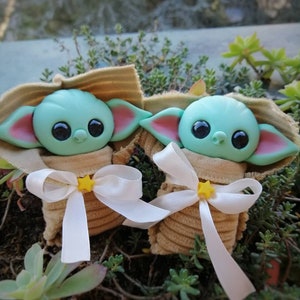 Puede incluir: Dos peque&ntilde;os juguetes de peluche verde y azul con forma de personaje de beb&eacute; alien&iacute;gena. Cada juguete est&aacute; envuelto en una tela marr&oacute;n con una cinta blanca y una estrella amarilla.