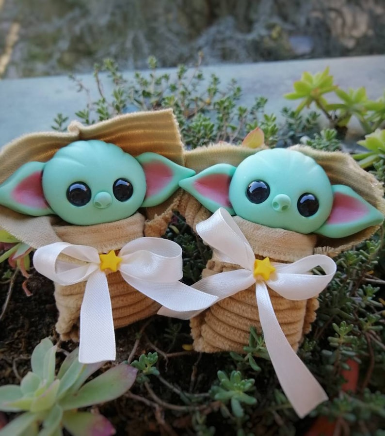 Puede incluir: Dos peque&ntilde;os juguetes de peluche verde y azul con forma de personaje de beb&eacute; alien&iacute;gena. Cada juguete est&aacute; envuelto en una tela marr&oacute;n con una cinta blanca y una estrella amarilla.