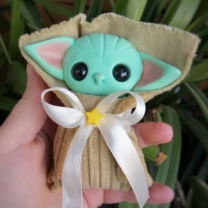Puede incluir: Un peque&ntilde;o juguete de peluche verde con ojos grandes y orejas puntiagudas, vestido con una bata de pana marr&oacute;n con una cinta de sat&eacute;n blanca y una estrella amarilla.