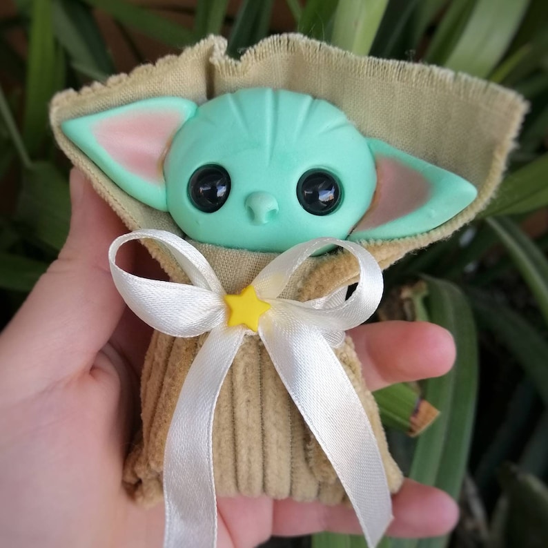 Puede incluir: Un peque&ntilde;o juguete de peluche verde con ojos grandes y orejas puntiagudas, vestido con una bata de pana marr&oacute;n con una cinta de sat&eacute;n blanca y una estrella amarilla.