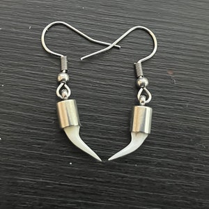 May include: A pair of silver earrings with white fang-shaped charms. The earrings have a simple hook design.