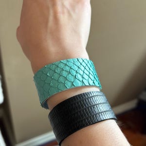May include: Two leather bracelets are shown. The top bracelet is a vibrant turquoise with a snakeskin pattern. The bottom bracelet is black with a textured, rectangular pattern. Both bracelets are wide cuff styles.
