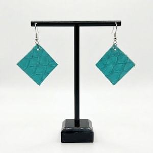 May include: A pair of turquoise leather earrings with a textured, scale-like pattern. The earrings are square-shaped and hang from silver-colored hooks. They are displayed on a black earring stand against a white background.