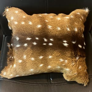 May include: A brown and white spotted deer hide pillow. The pillow is made from real deer hide and has a soft, plush texture.