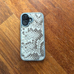 May include: A smartphone case with a snakeskin pattern in shades of gray and white. The case has a light blue border and a cutout for the camera lenses. The case is lying on a wooden surface.