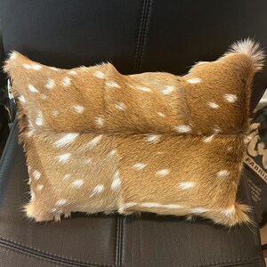 May include: A brown and white spotted deer hide pillow. The pillow is rectangular and has a soft, fuzzy texture.