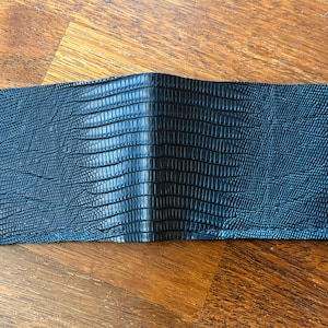 May include: A black bi-fold wallet with a textured, reptile-like pattern. The wallet is made of a dark material and is open, revealing the interior. The wallet is rectangular and appears to be made of leather.