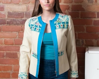May include: A cream-coloured long-sleeve jacket featuring a bright turquoise zip trim and collarless neckline. The garment is accented with embroidered panels on the shoulders, pockets, and cuffs, displaying geometric patterns in red, blue, and green.