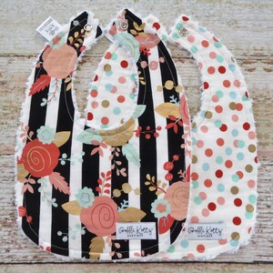 May include: Three baby bibs with different patterns. One bib has black and white stripes with a floral design in pink, gold, and green. Another bib has a white background with red, gold, and blue polka dots. The third bib has a white background with red, gold, and blue polka dots. All bibs have a white ruffled edge and a label that says "Ruffle Kottz Boutique".