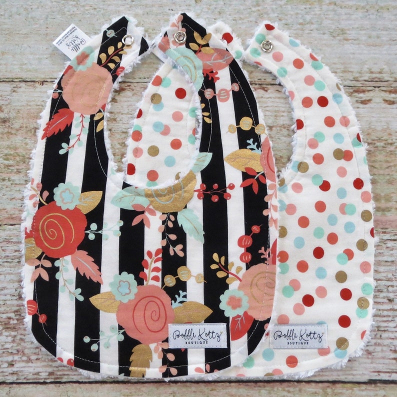May include: Three baby bibs with different patterns. One bib has black and white stripes with a floral design in pink, gold, and green. Another bib has a white background with red, gold, and blue polka dots. The third bib has a white background with red, gold, and blue polka dots. All bibs have a white ruffled edge and a label that says "Ruffle Kottz Boutique".