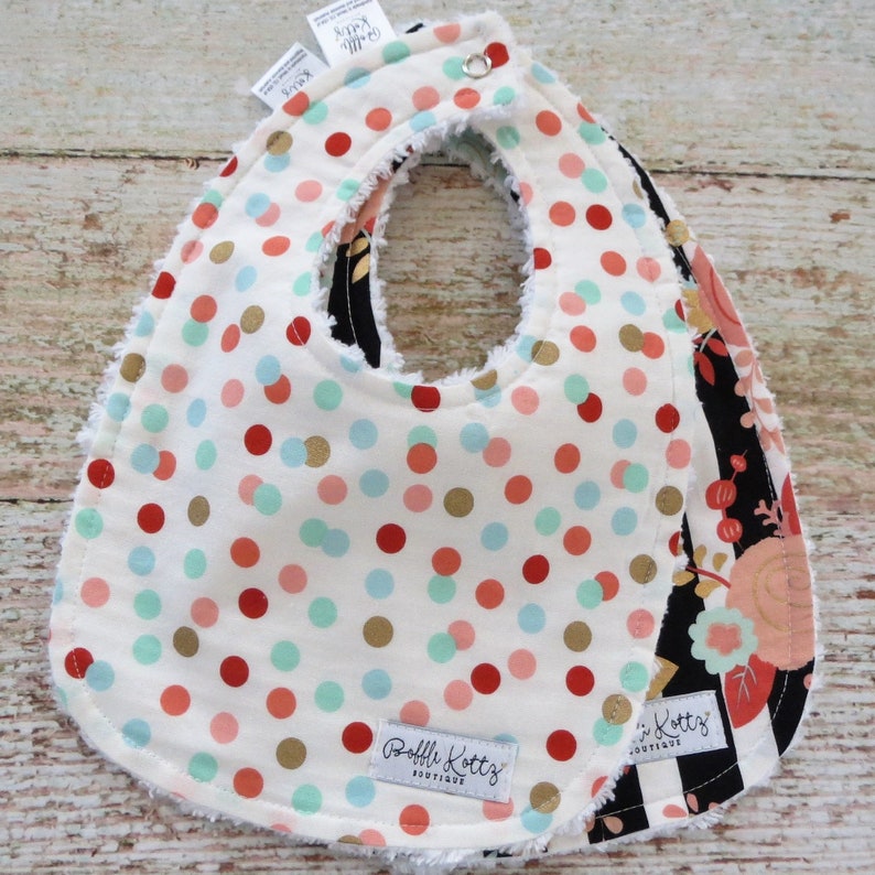 May include: Two-layer baby bib with a white background and red, gold, and blue polka dots. The top layer has a white background with red, gold, and blue polka dots. The bottom layer has a black background with a floral pattern in pink, gold, and white. The bib has a ruffled edge and a snap closure. The bib is labeled "Ruffle Kottz Boutique".