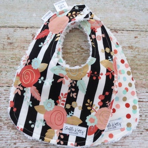 May include: A black and white striped bib with a floral pattern and gold accents. The bib has a white fleece backing and a red polka dot pattern on the back. The bib has a snap closure and the text "Bobble Kotty's Boutique" is printed on the front.