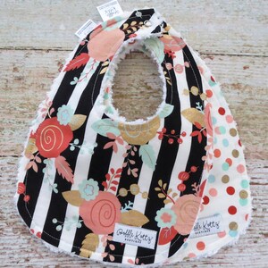 May include: A black and white striped baby bib with a floral pattern and gold accents. The bib has a white fleece lining and a snap closure. The bib is reversible and has a polka dot pattern on the other side. The bib has the text "Raffili Kottz Boutique" on it.