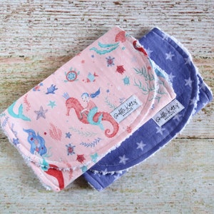 May include: Two baby burp cloths. One is pink with a mermaid and seahorse design, the other is blue with white stars. Both have a white tag that says "Bobbi Kottz Boutique".