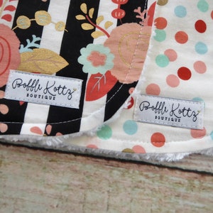 May include: Two white fabric baby bibs with black and white stripes and polka dots. The bibs have a white label with the text "Bobbli Kottz Boutique" on them.