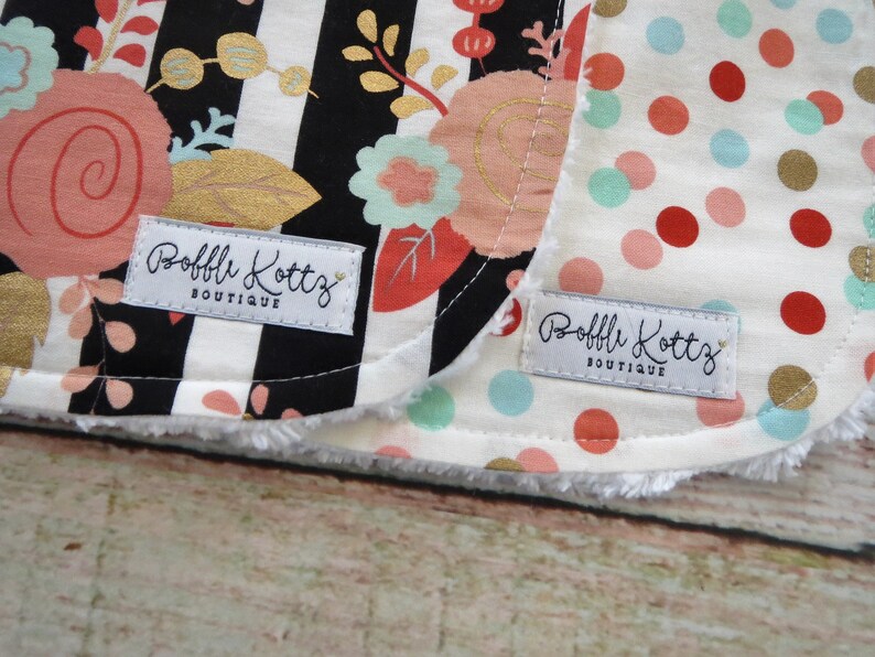 May include: Two white fabric baby bibs with black and white stripes and polka dots. The bibs have a white label with the text "Bobbli Kottz Boutique" on them.