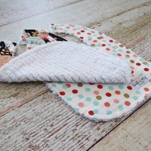 May include: Two baby bibs, one with a white terry cloth backing and a colorful polka dot print, and the other with a floral print. The polka dot bib has a gold, red, and turquoise polka dot pattern on a white background. The floral bib has a black background with pink, green, and gold floral accents. Both bibs have a white terry cloth backing.