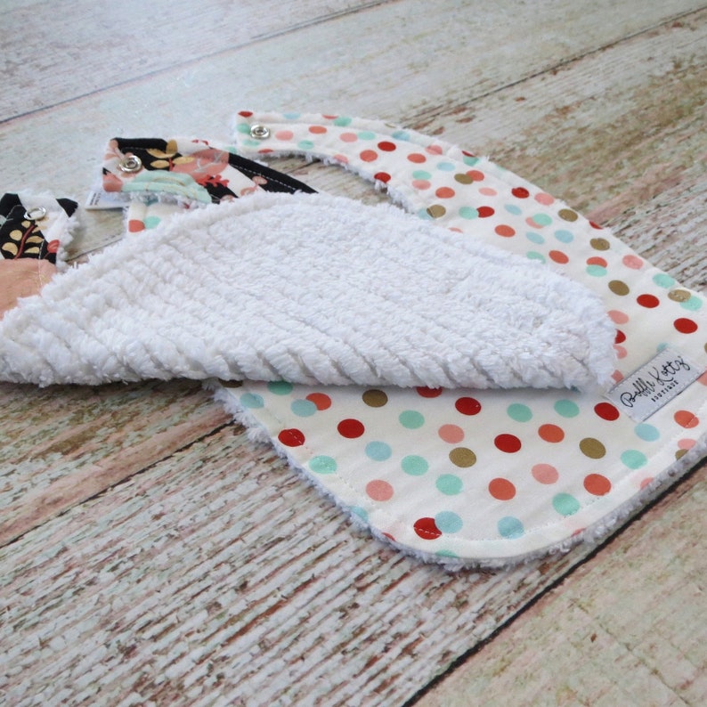 May include: Two baby bibs, one with a white terry cloth backing and a colorful polka dot print, and the other with a floral print. The polka dot bib has a gold, red, and turquoise polka dot pattern on a white background. The floral bib has a black background with pink, green, and gold floral accents. Both bibs have a white terry cloth backing.