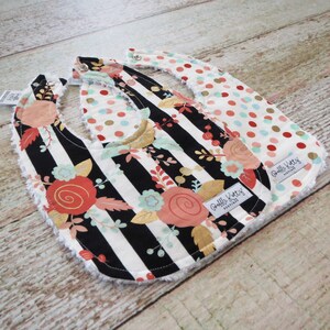 May include: Two reversible baby bibs with a black and white striped pattern with gold accents and floral designs on one side and a red, white, and gold polka dot pattern on the other side. The bibs have a soft, white terry cloth backing and are labeled "Ruffle Buttz Boutique".