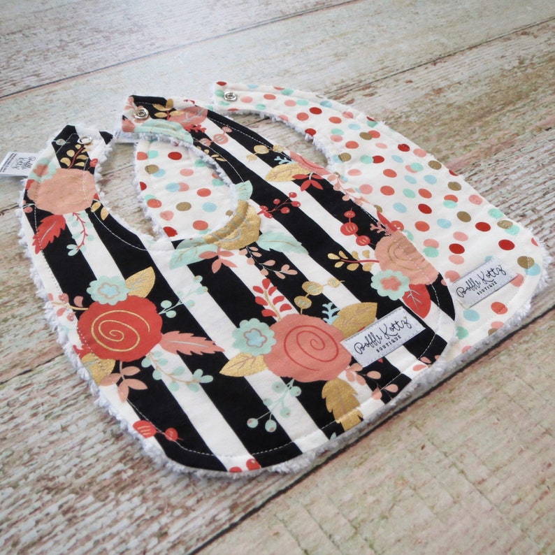May include: Two reversible baby bibs with a black and white striped pattern with gold accents and floral designs on one side and a red, white, and gold polka dot pattern on the other side. The bibs have a soft, white terry cloth backing and are labeled "Ruffle Buttz Boutique".